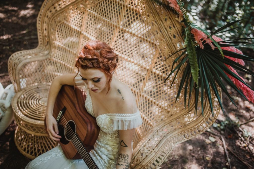 redhead bride sitting in rattan peacock chair playing acoustic guitar