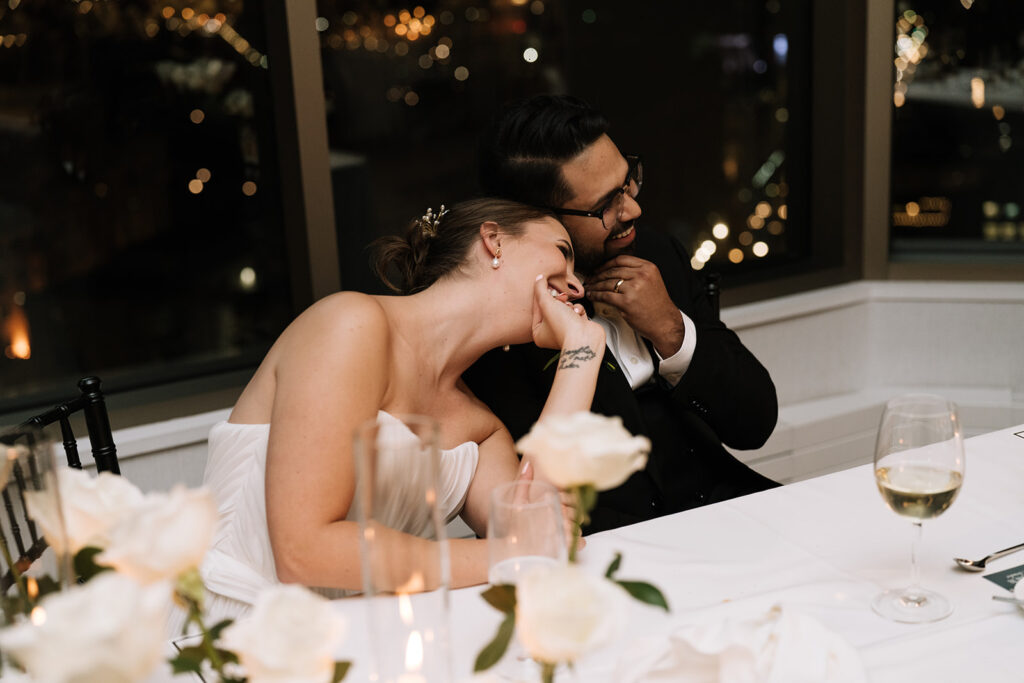 Bride and groom share a tender moment being present at their reception table overlooking city lights, photographed by Danielle Aisling.
